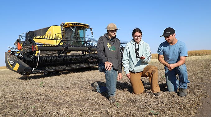 A group of students kneel in front of a combine and inspect some wheat during Harvest 2023