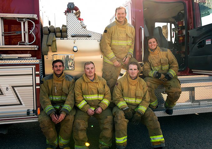 A group of Lakeland College firefighting students dressed in gear sit together along the side of a fire truck.