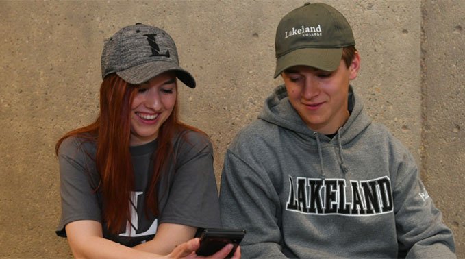 Two students wearing Lakeland College hats and sweaters look at a cell phone