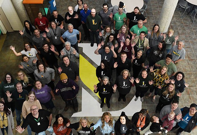 Lakeland College employees, standing around a superimposed image of the college's L logo, wave up to the camera