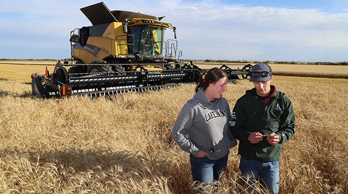 A combine sis parked behind a couple of crop technology students who inspect a strand of wheat. 