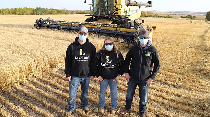Three students wearing Lakeland College sweaters stand in front of a New Holland combine in a crop field.