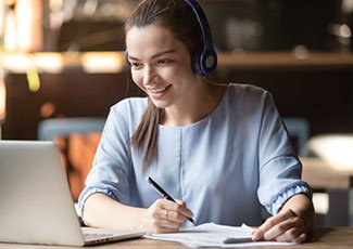 female sitting in front of laptop, wearing headphones while taking notes in notebook