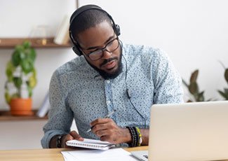male sitting at laptop, wearing headphones while taking notes in a notebook